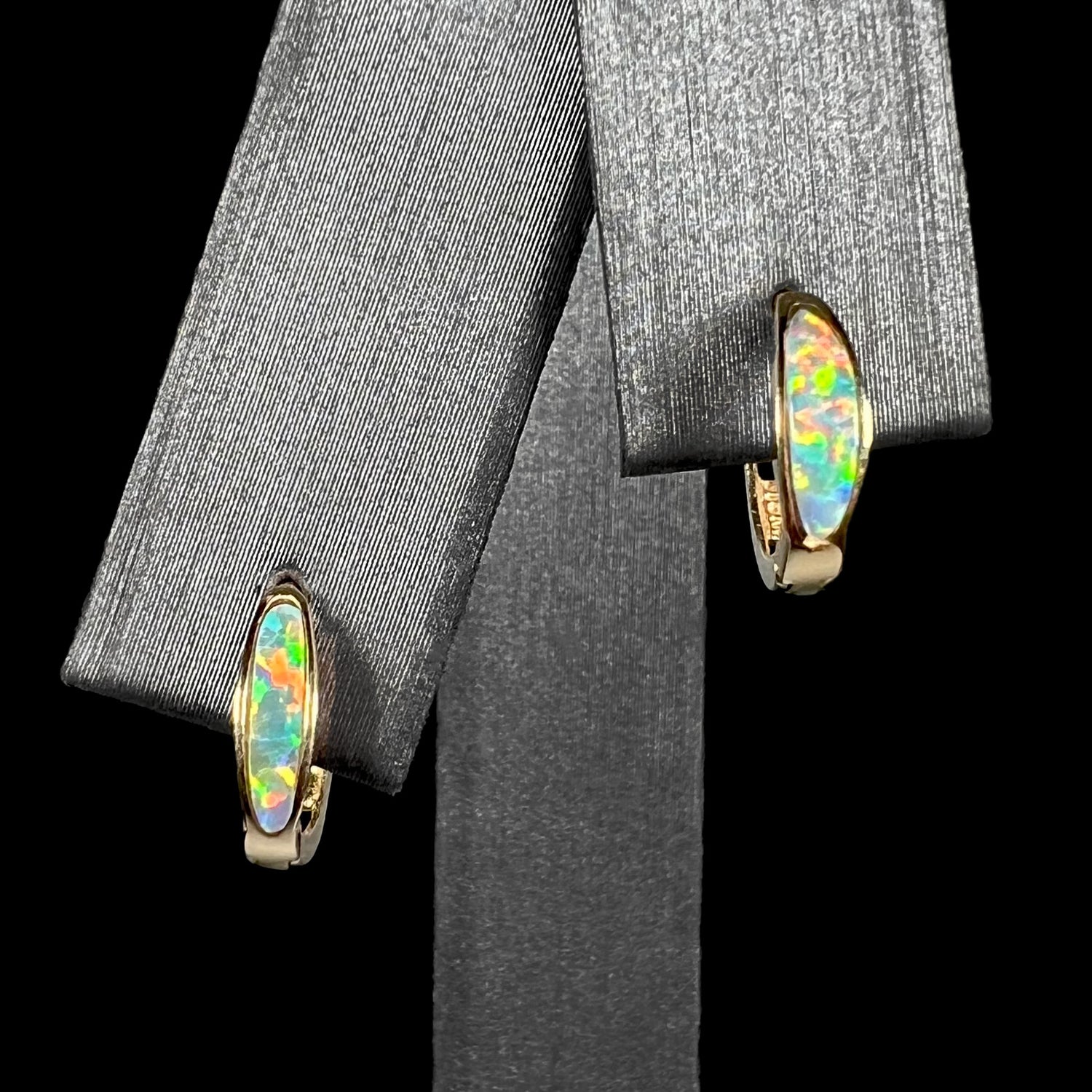 A pair of yellow gold lab-created opal inlay huggie hoop earrings shown on an earring display against a black background.