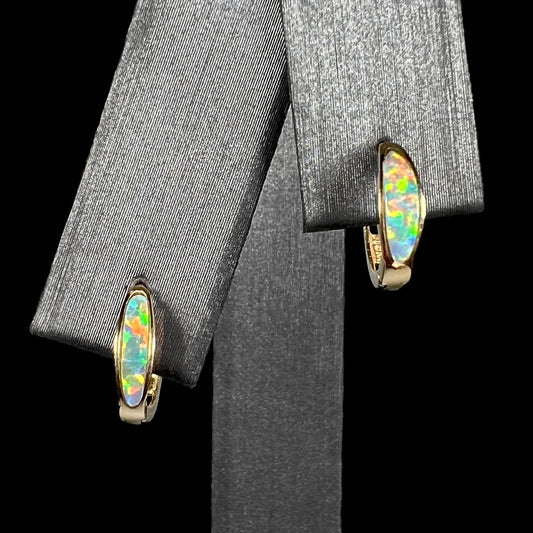 A pair of yellow gold lab-created opal inlay huggie hoop earrings shown on an earring display against a black background.