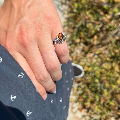 A ladies' pear shaped red fire agate ring with a sterling silver leaf design worn on a hand's pinky finger as it rests on the model's hip.