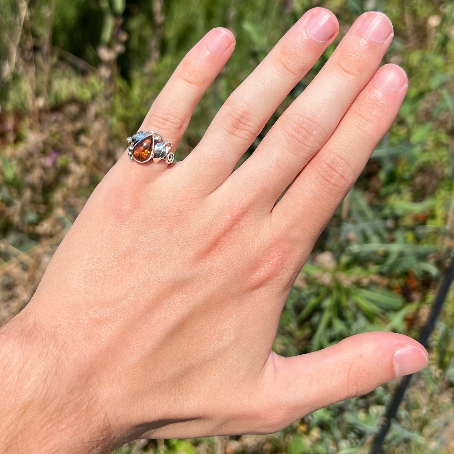 A ladies' pear shaped red fire agate ring with a sterling silver leaf design worn on a hand's pinky finger in sunlight.