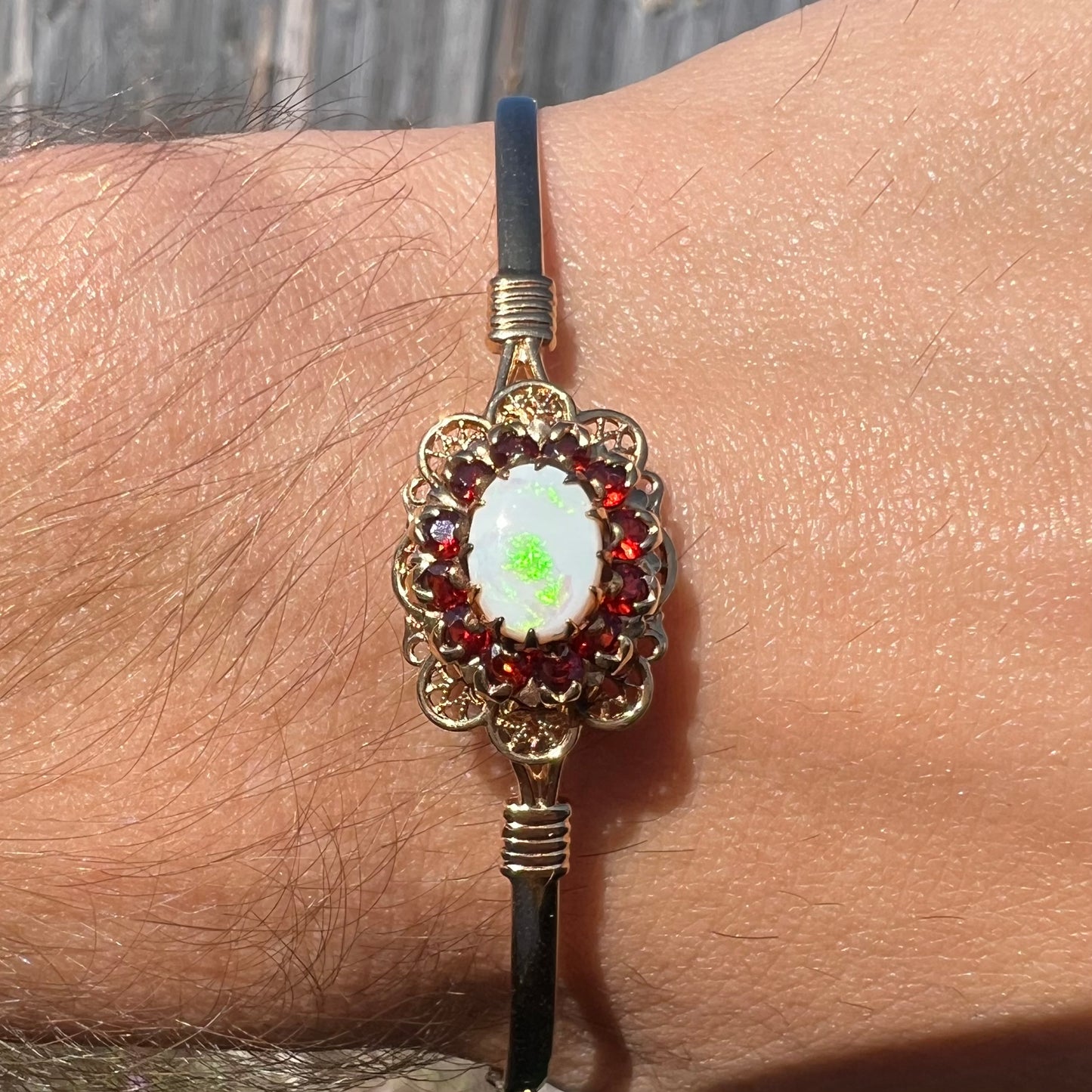A vintage yellow gold Australian opal and garnet halo bangle bracelet shown on a wrist close-up in sunlight.