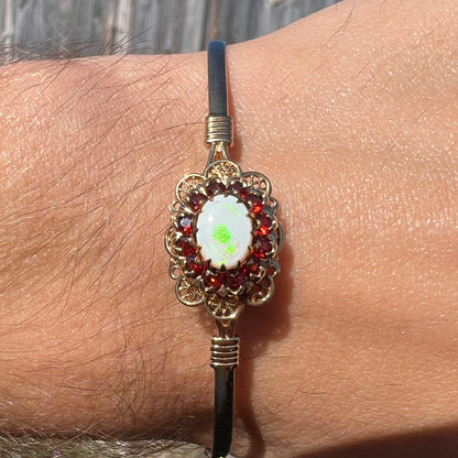 A vintage yellow gold Australian opal and garnet halo bangle bracelet shown on a wrist close-up in sunlight.