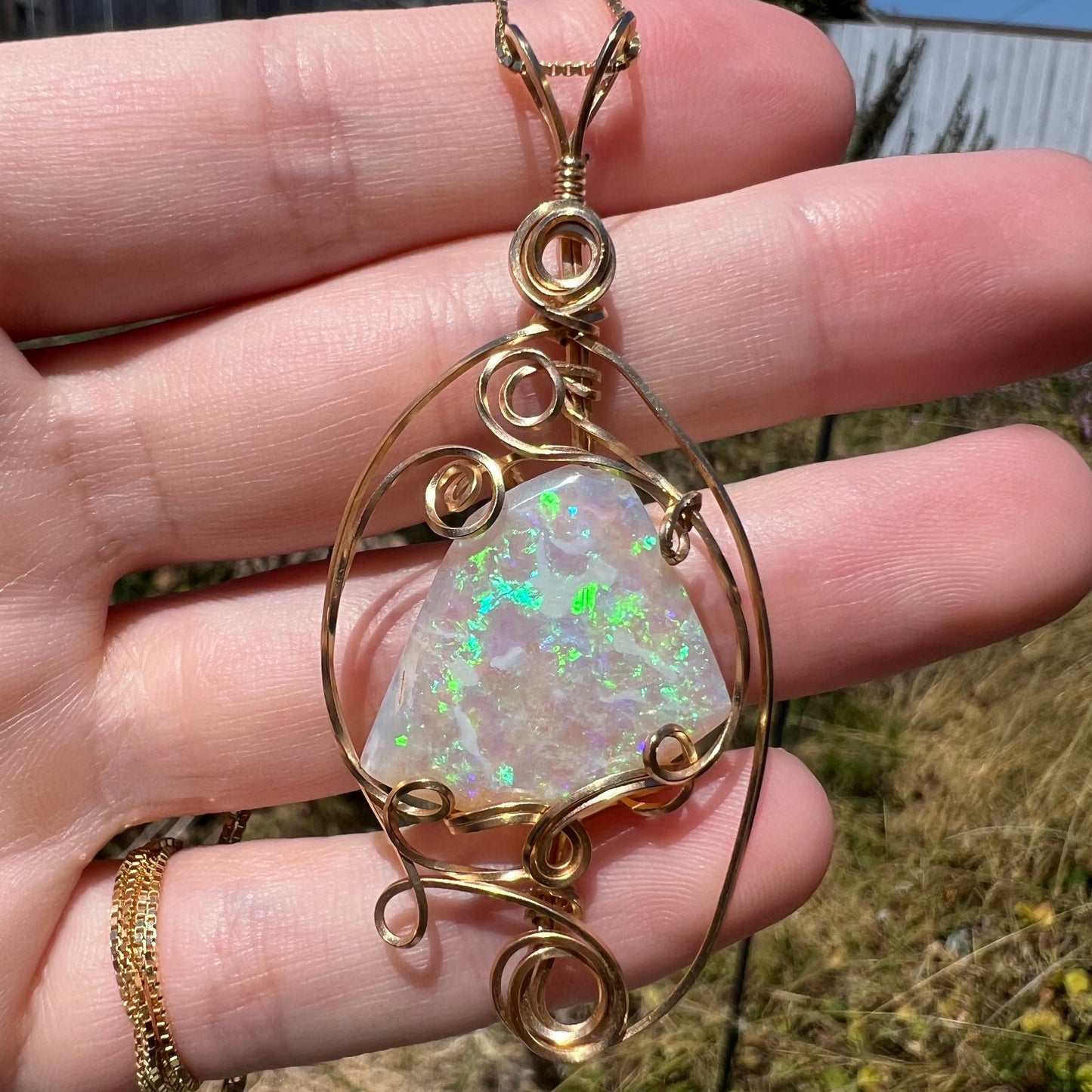 Close-up sunlit view of a Lambina, Australian crystal opal pendant with green and blue flashes of color, set in a gold-tone wire wrapped mounting.