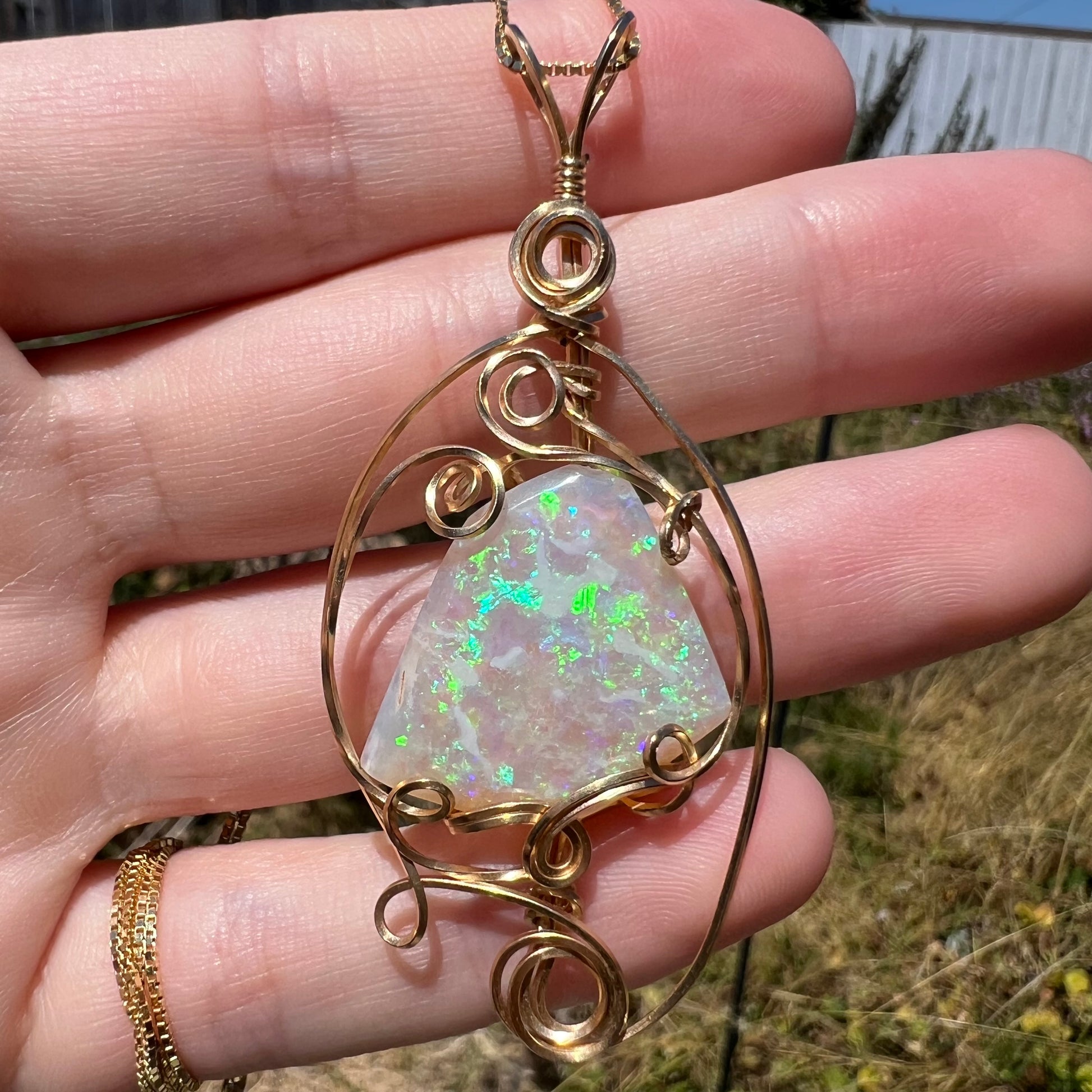 Close-up sunlit view of a Lambina, Australian crystal opal pendant with green and blue flashes of color, set in a gold-tone wire wrapped mounting.
