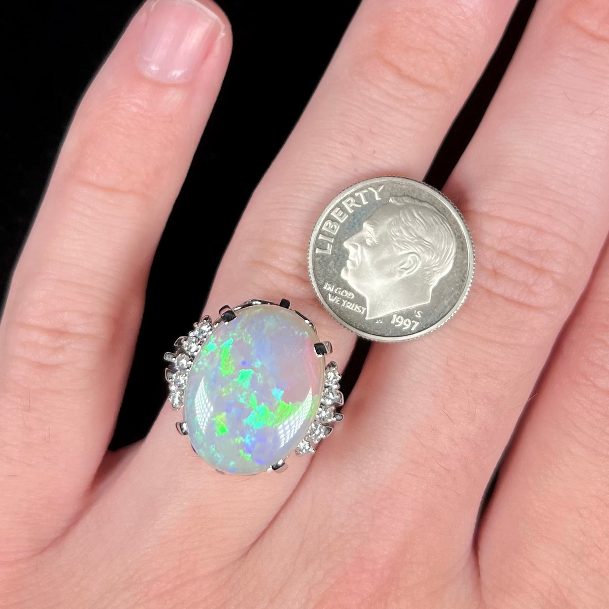 Close-up of hand wearing a large, oval-shaped Australian crystal opal and diamond cocktail ring next to US dime coin for scale