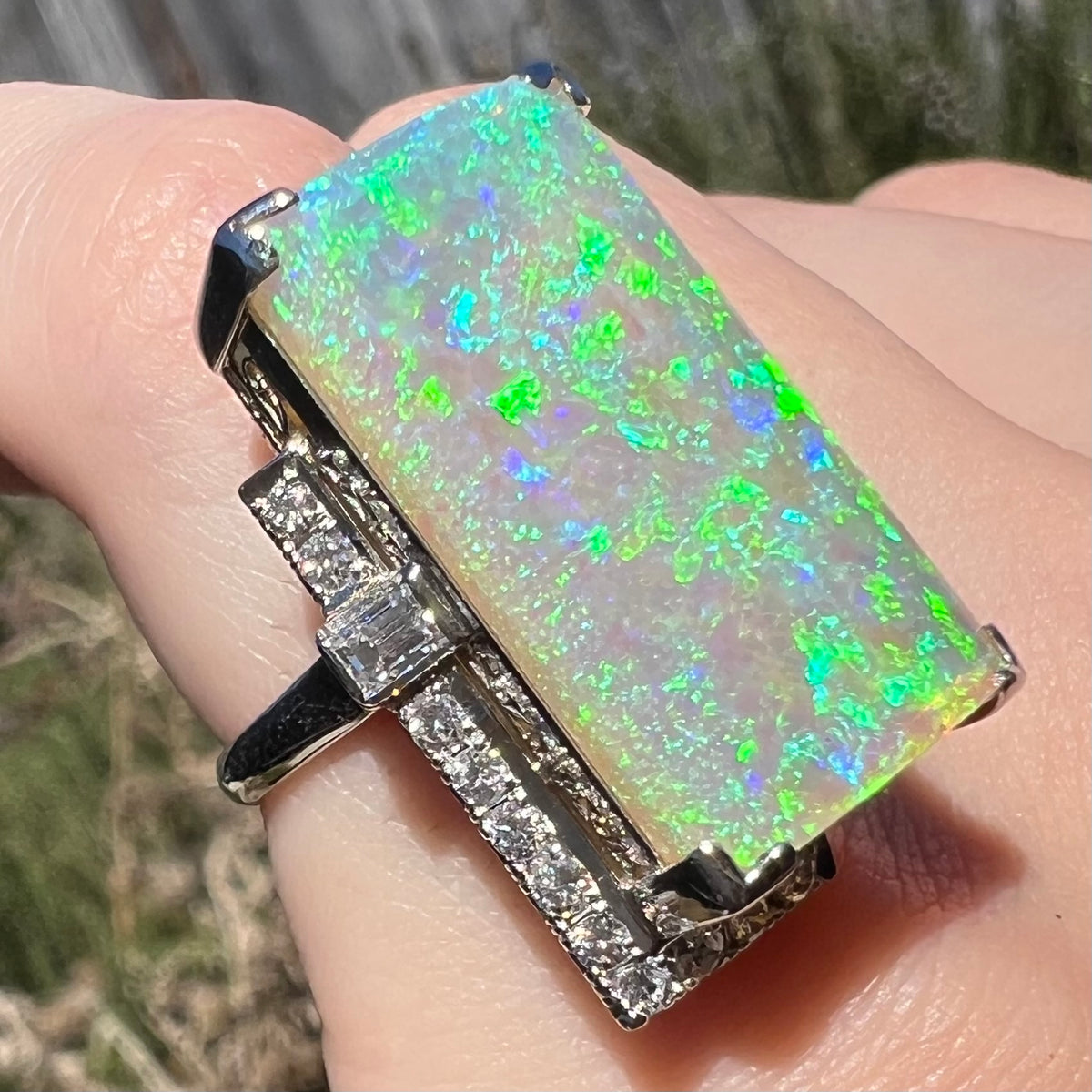 Green and blue play-of-color is seen across the broad surface of a large rectangular Australian opal and diamond cocktail ring, worn on finger in sunlight.