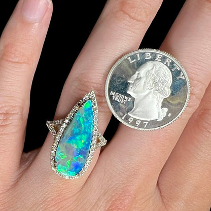 A large, blue, pear-shaped boulder opal and diamond ring worn on a hand next to a quarter coin for scale.