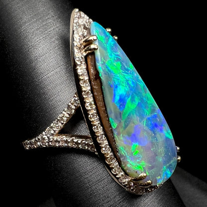 Angled close-up of a large boulder opal and diamond halo engagement ring with a split shank design, shown on a ring display.