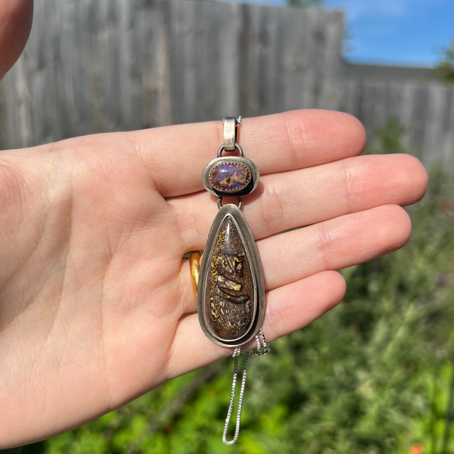 A large two-stone boulder opal necklace in sterling silver held in a hand under natural sunlight against a blurred outdoor backdrop.
