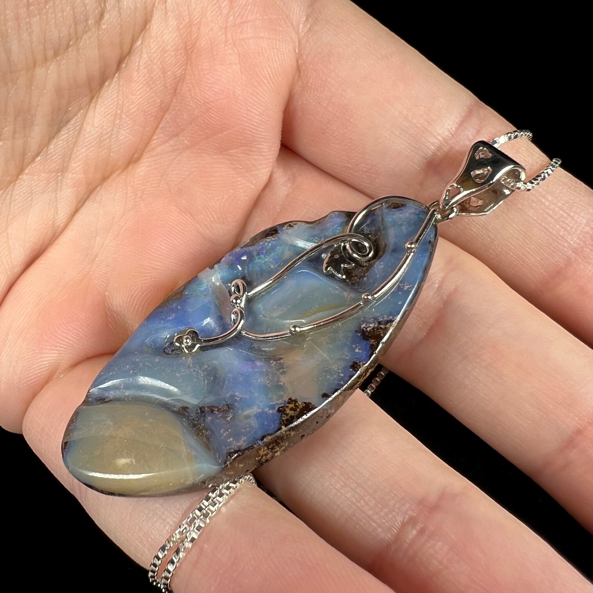 Angled close-up of a large, blue Australian boulder opal necklace in sterling silver, shown in a hand.