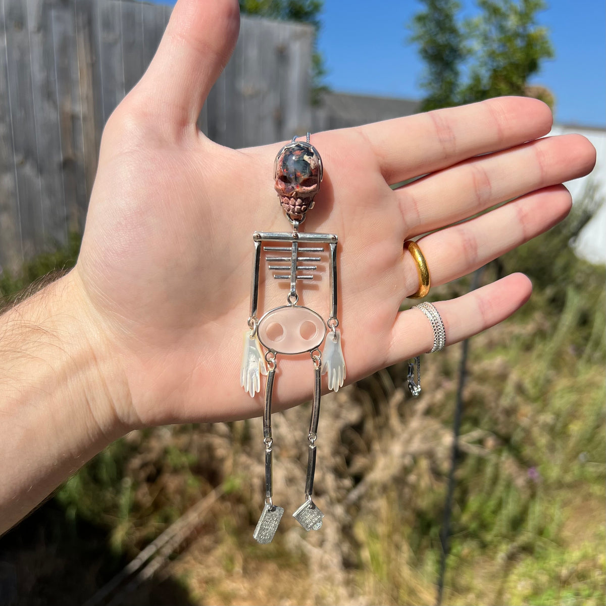 Hand holding a large "dancing" articulated skeleton pendant made from sterling silver, 14k gold, Mexican cantera opal, and mother of pearl, held in sunlight