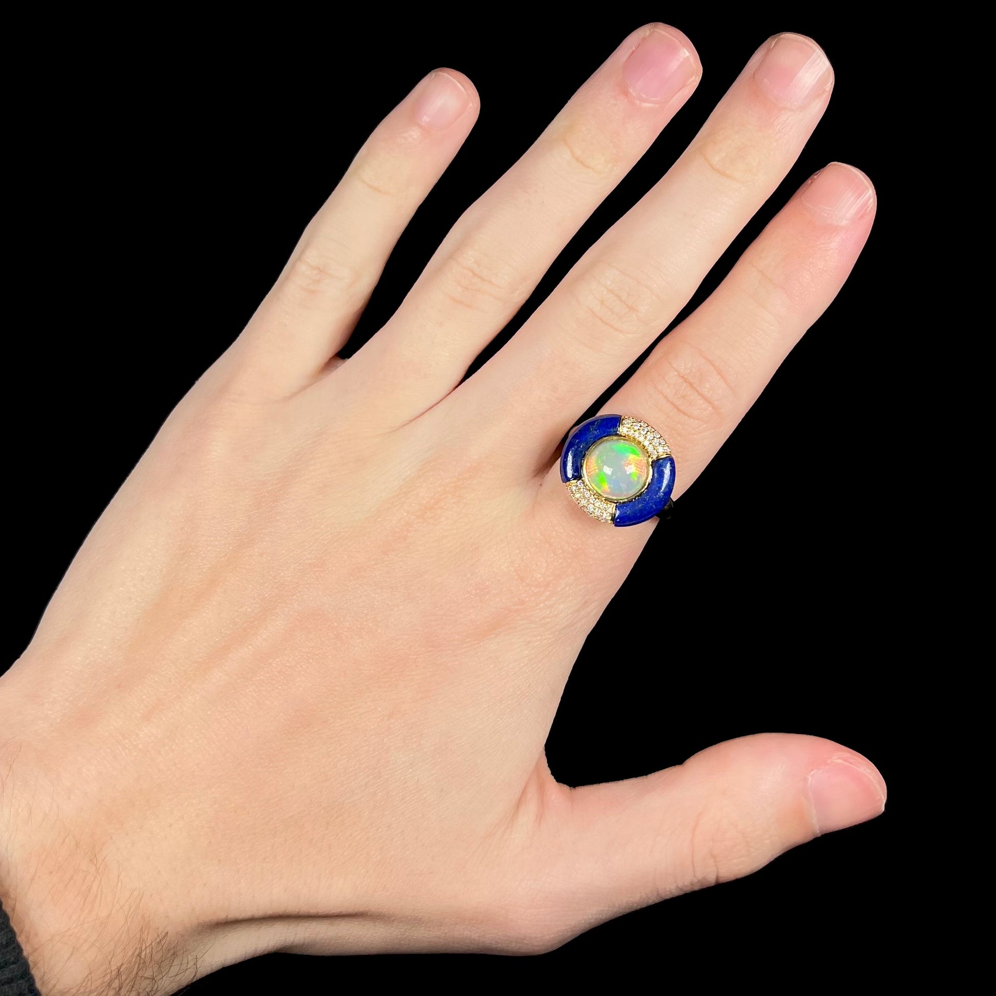 A large women's Ethiopian opal, lapis lazuli, and diamond ring in 14k gold by Cirari, worn on the index finger of a hand against a black background.