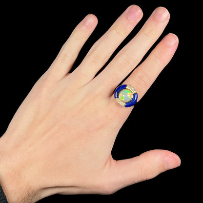 A large women's Ethiopian opal, lapis lazuli, and diamond ring in 14k gold by Cirari, worn on the index finger of a hand against a black background.