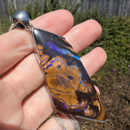 Angled close-up of large, freeform cut boulder opal pendant with veins of precious opal, held in hand under natural light