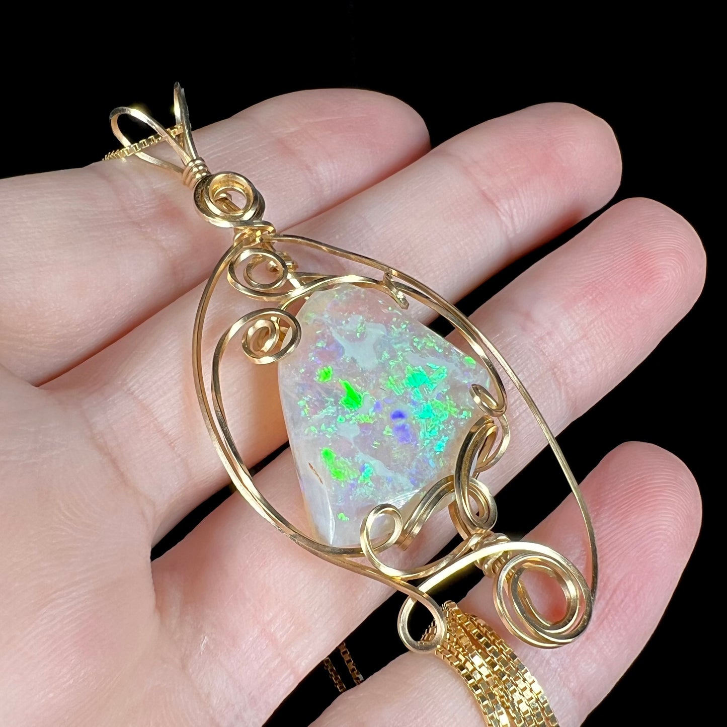 Angled close-up of a large crystal opal pendant with green and blue play-of-color featuring a gold-plated wire wrapped design, resting on fingers.