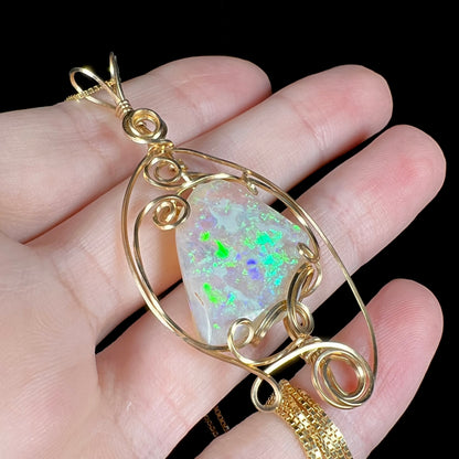 Angled close-up of a large crystal opal pendant with green and blue play-of-color featuring a gold-plated wire wrapped design, resting on fingers.