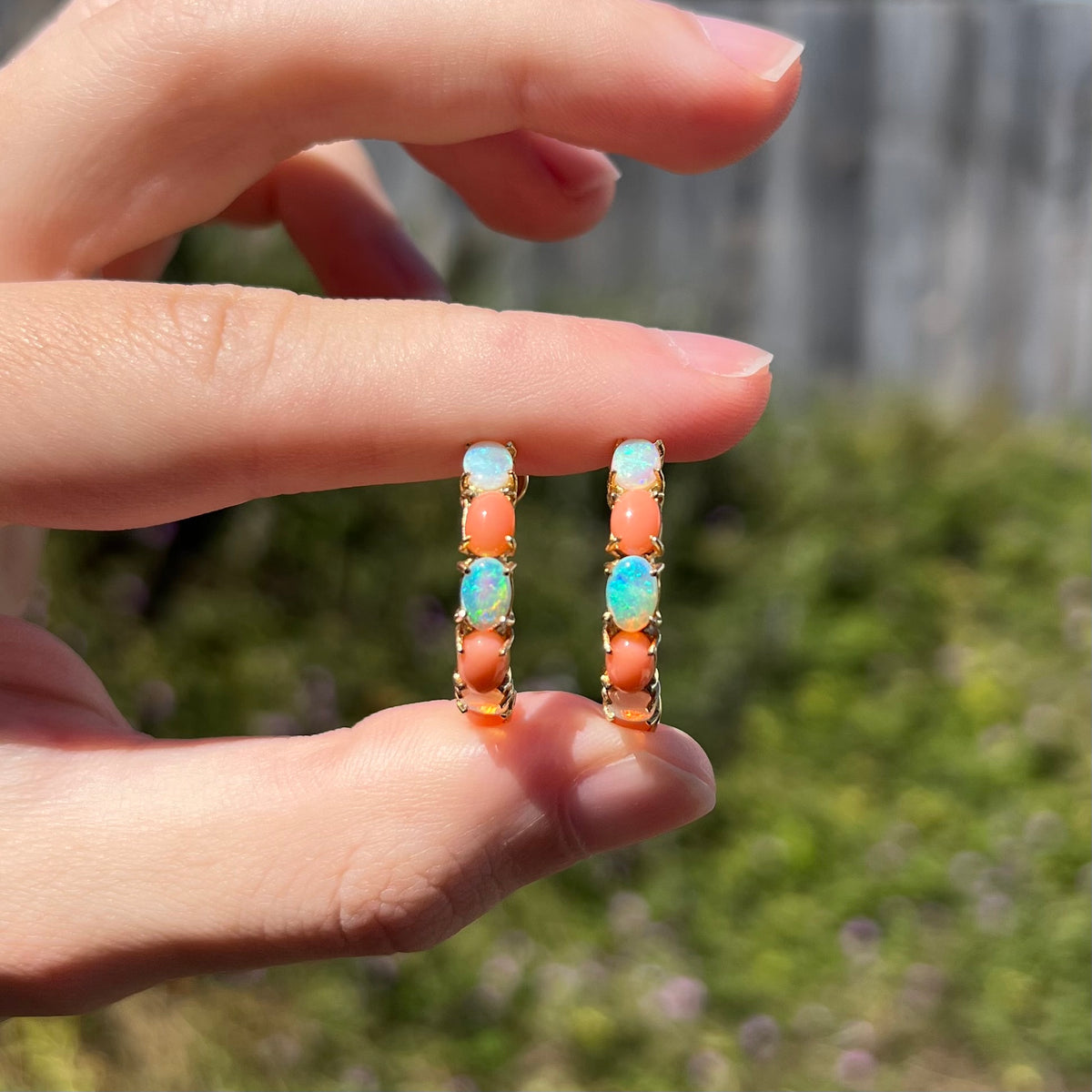 Two fingers holding pair of large, prong-set Australian crystal opal and coral semi-hoop earrings in sunlight