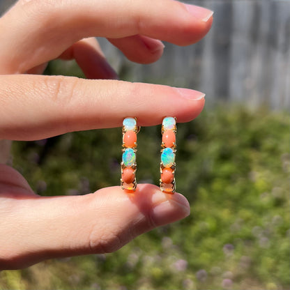 Two fingers holding pair of large, prong-set Australian crystal opal and coral semi-hoop earrings in sunlight
