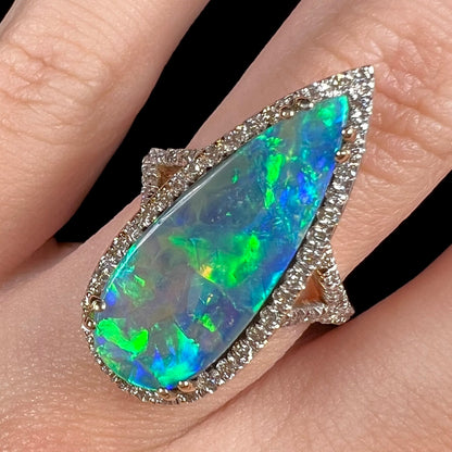 A large, elongated pear-shaped boulder opal and diamond halo ring shown close-up on a finger.