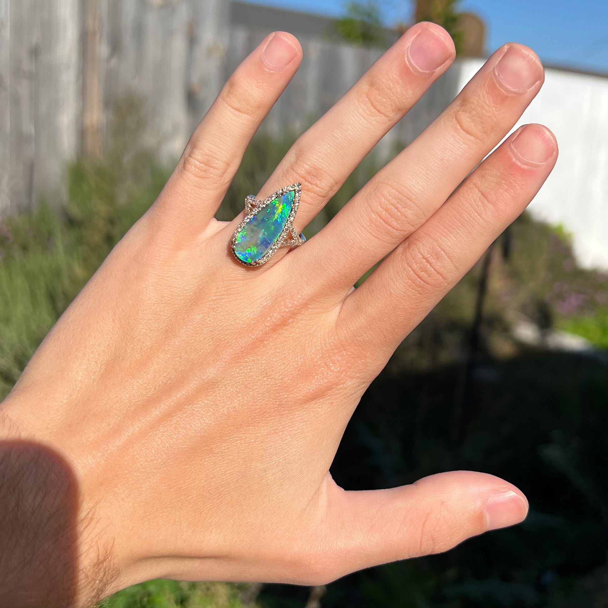 Hand wearing a large, pear-shaped boulder opal and diamond pave halo ring in the sunlight against blurred outdoor background.