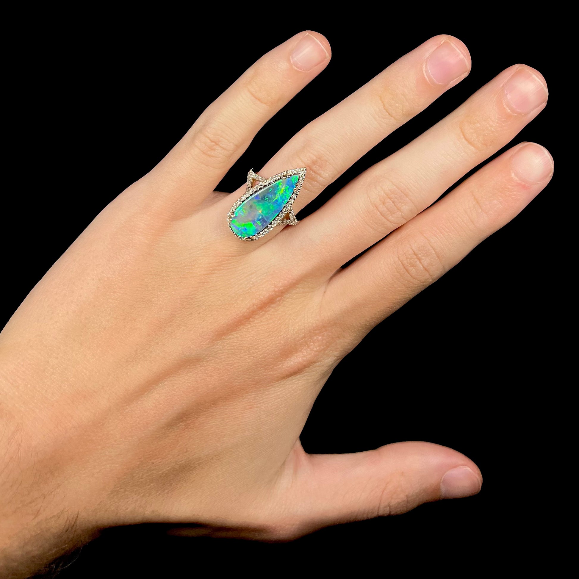 Hand wearing a large pear shaped boulder opal and diamond engagement ring with green color play, shown against a black background.
