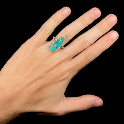 Hand wearing a large pear shaped boulder opal and diamond engagement ring with green color play, shown against a black background.