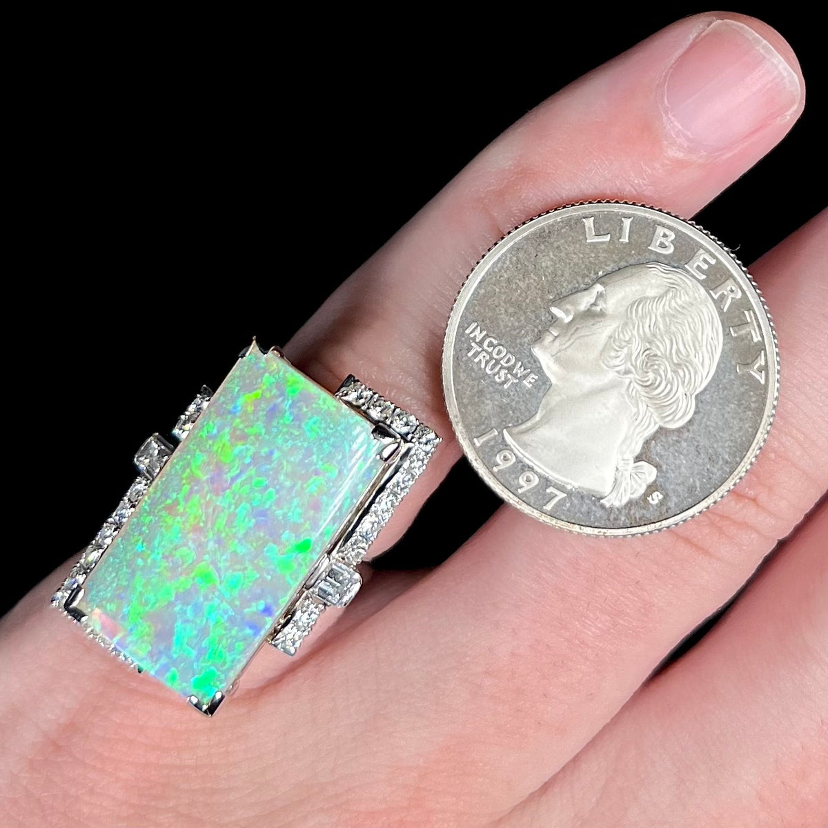 Close-up of a large, rectangular 11.46ct Australian opal and diamond cocktail ring worn on hand next to US quarter coin for scale.