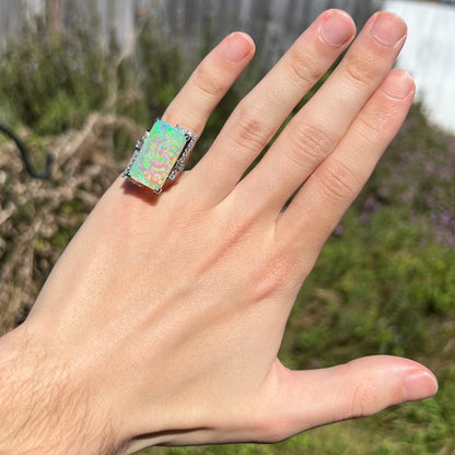 Large, rectangular Australian opal and diamond pinky ring worn on hand in natural sunlight.  Green play-of-color is evenly distributed across the opal, a pinkish undertone the result of its semi-crystal body.