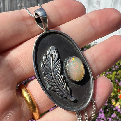 Angled close-up of a large sterling silver opal and feather design pendant held in a hand under natural sunlight.
