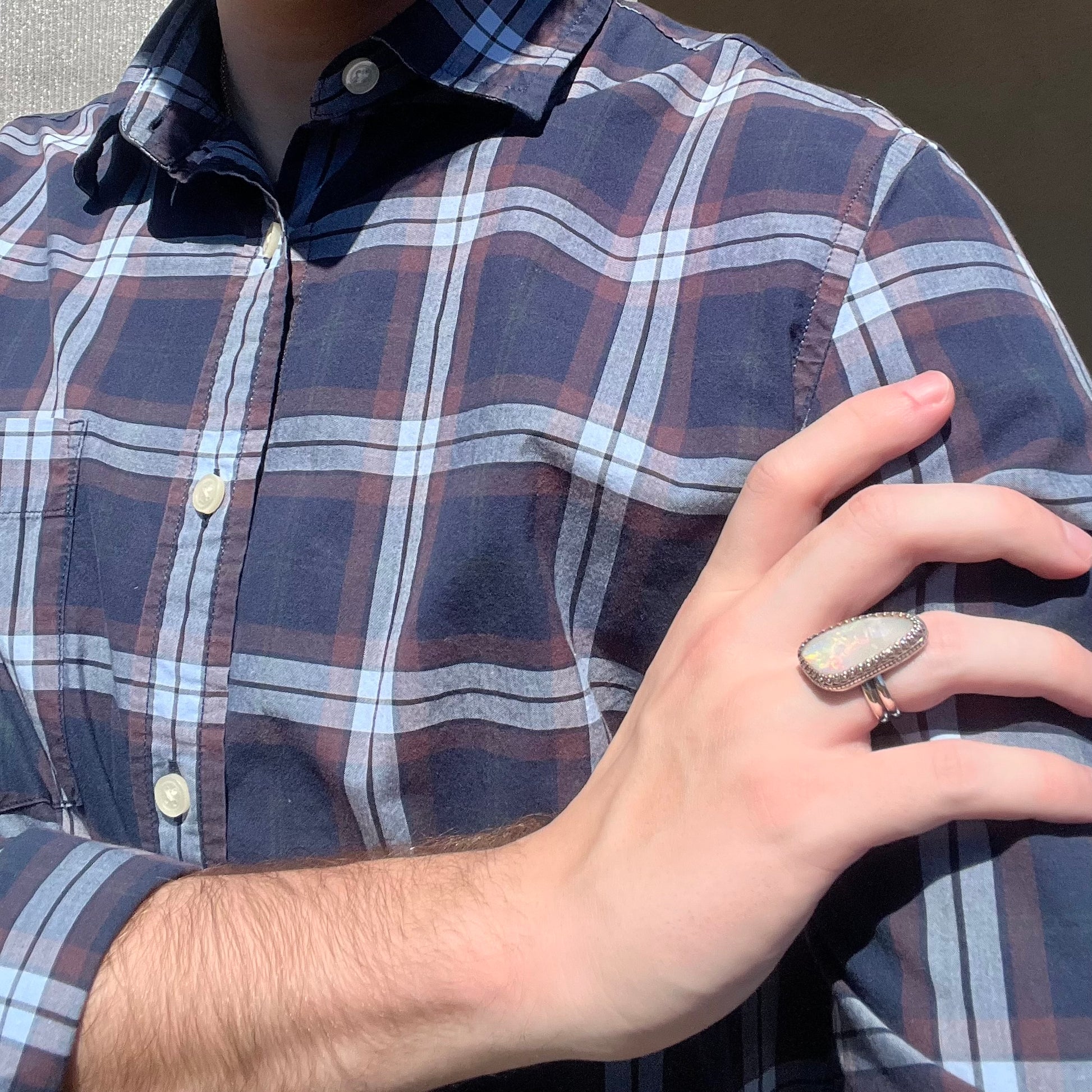 Hand resting on person's shoulder against blue plaid shirt wearing a large, unisex sterling silver opal ring in sunlight