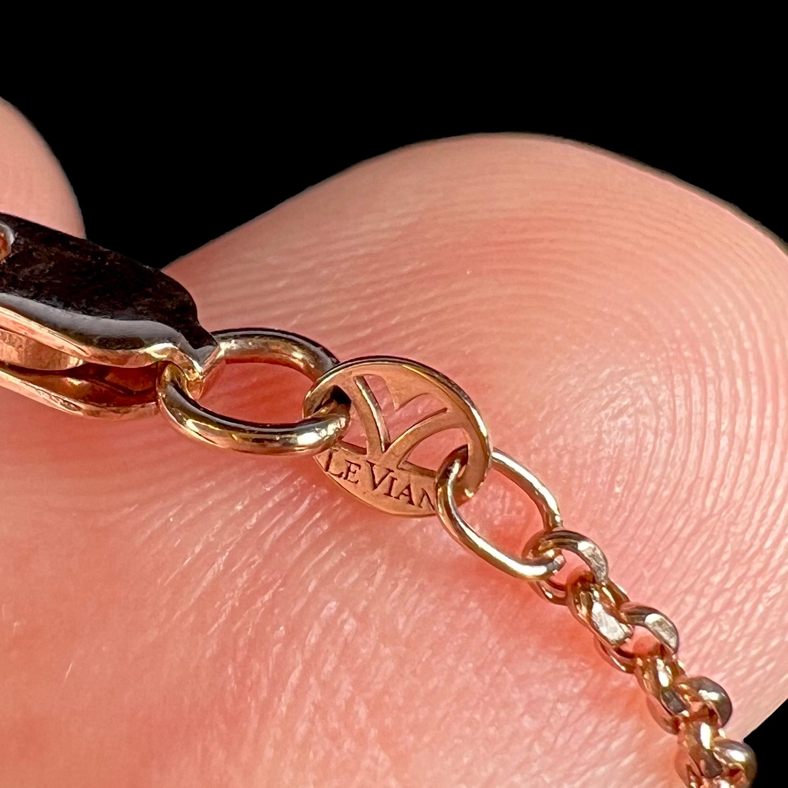 Close-up of a rose gold chain's hallmark tag stamped with the words "LE VIAN".  The tag rests on the tip of a finger against a black background.