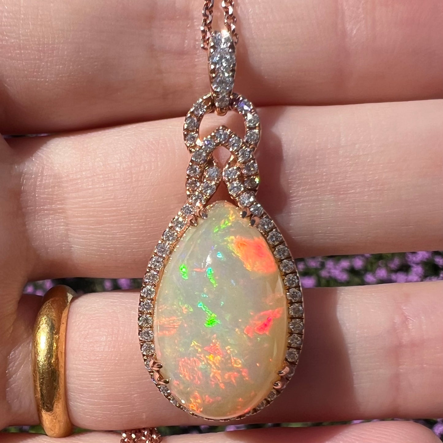 Close-up of an 18k rose gold opal and diamond halo necklace by Le Vian, shown held in a hand beneath natural sunlight.