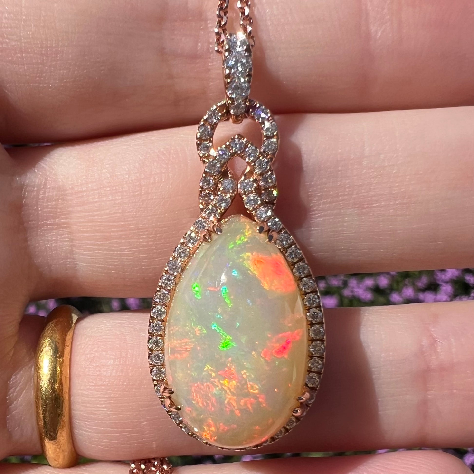 Close-up of an 18k rose gold opal and diamond halo necklace by Le Vian, shown held in a hand beneath natural sunlight.