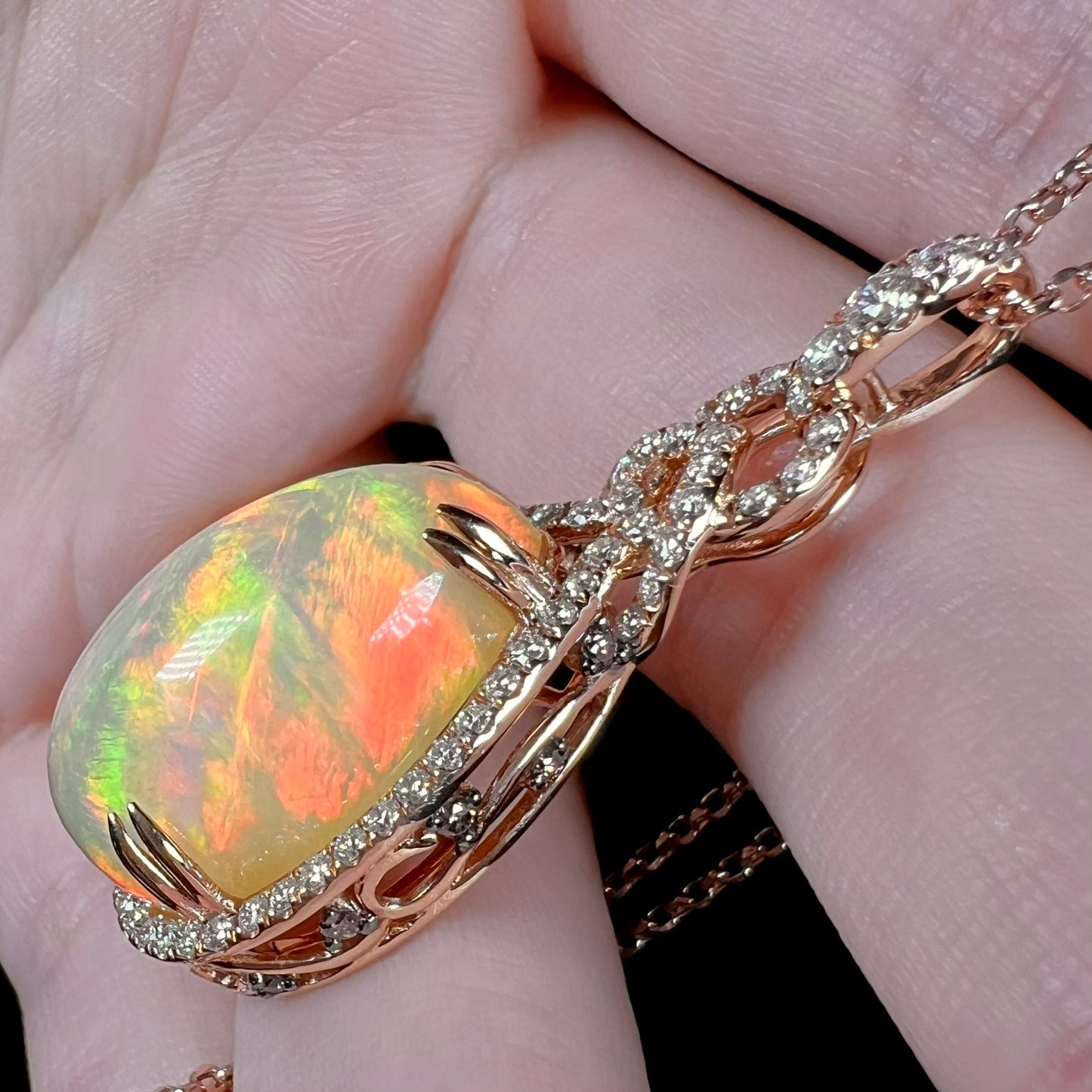 A rose gold opal and diamond necklace by Le Vian shown resting against fingers at an angle.  The opal displays red fire along its high dome.