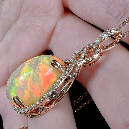A rose gold opal and diamond necklace by Le Vian shown resting against fingers at an angle.  The opal displays red fire along its high dome.