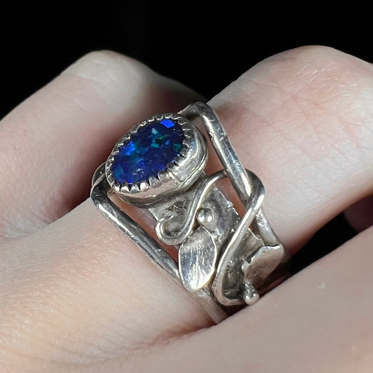 Angled close-up side view of scrolling leaf and vine design on sterling silver black opal doublet ring, worn on finger