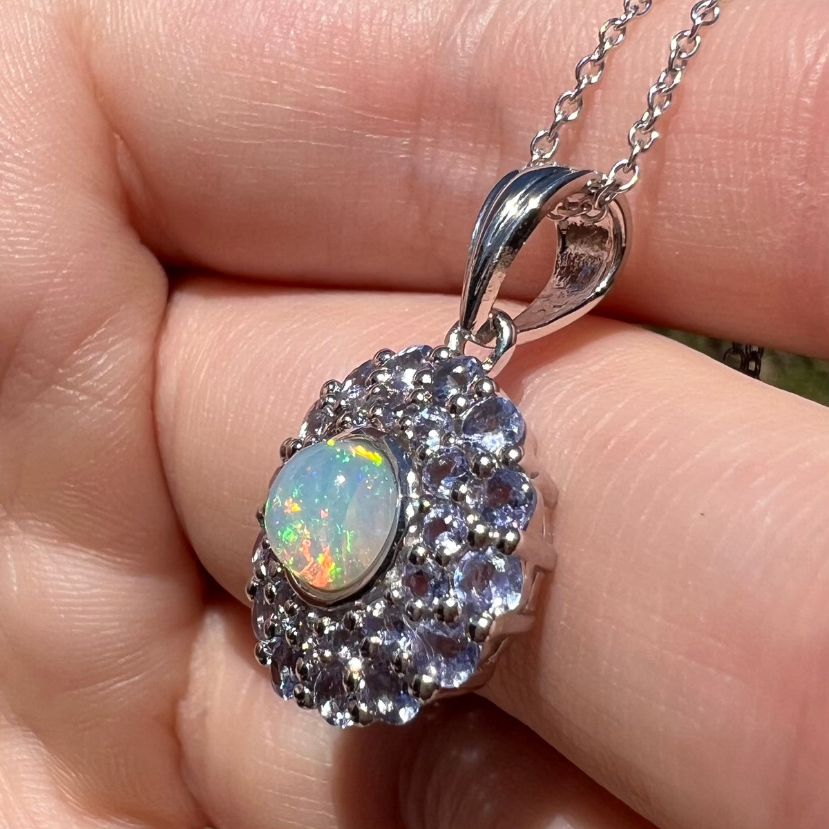 Angled close-up of a sterling silver opal pendant with a light blue tanzanite halo, shown in sunlight close-up.