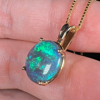 An oval shaped Lightning Ridge, Australian black opal pendant in 14k gold shown resting on fingers, close-up from angle
