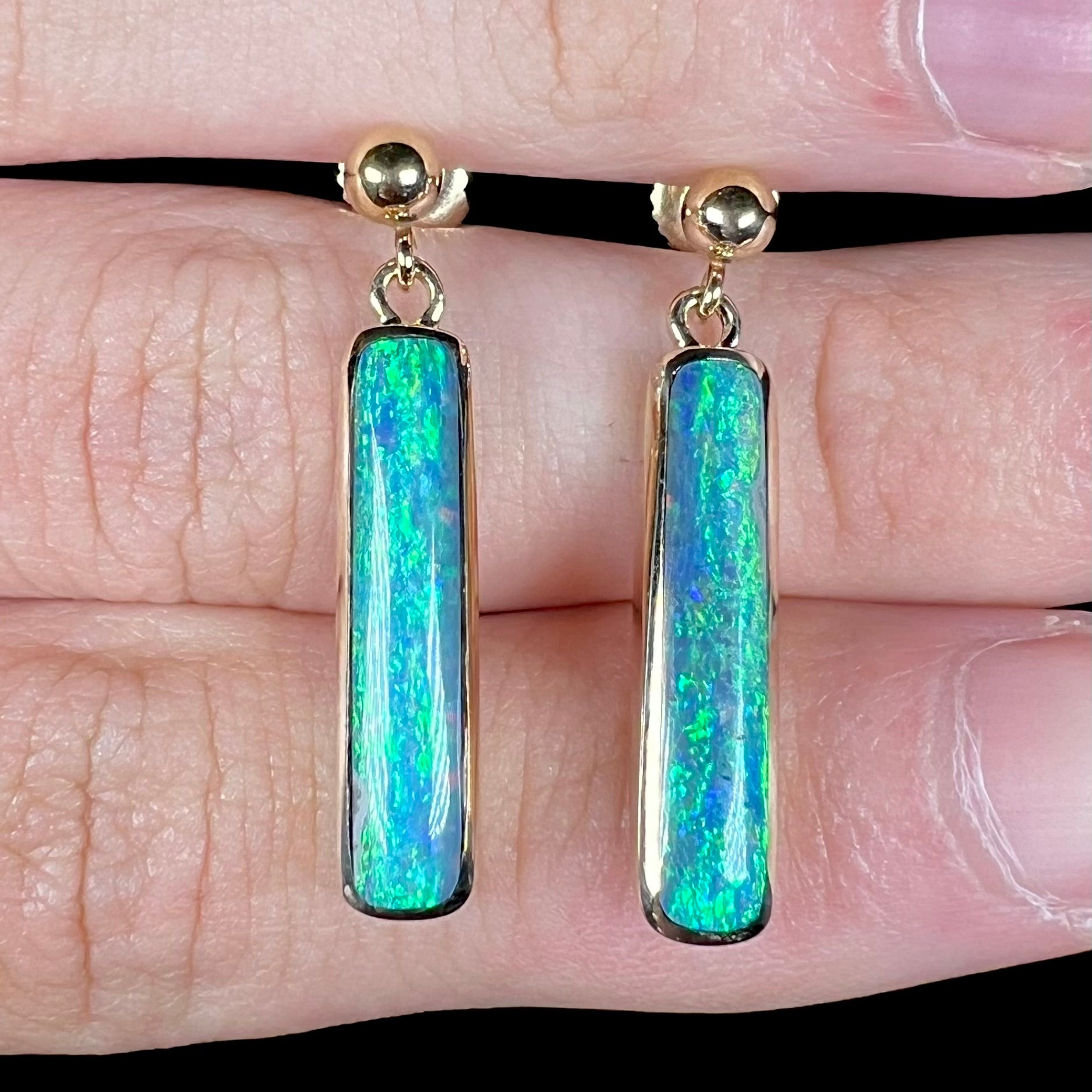 Close-up front view of a pair of Lightning Ridge opal dangle earrings held between two fingers on a hand.