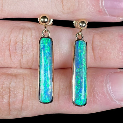 Close-up front view of a pair of Lightning Ridge opal dangle earrings held between two fingers on a hand.