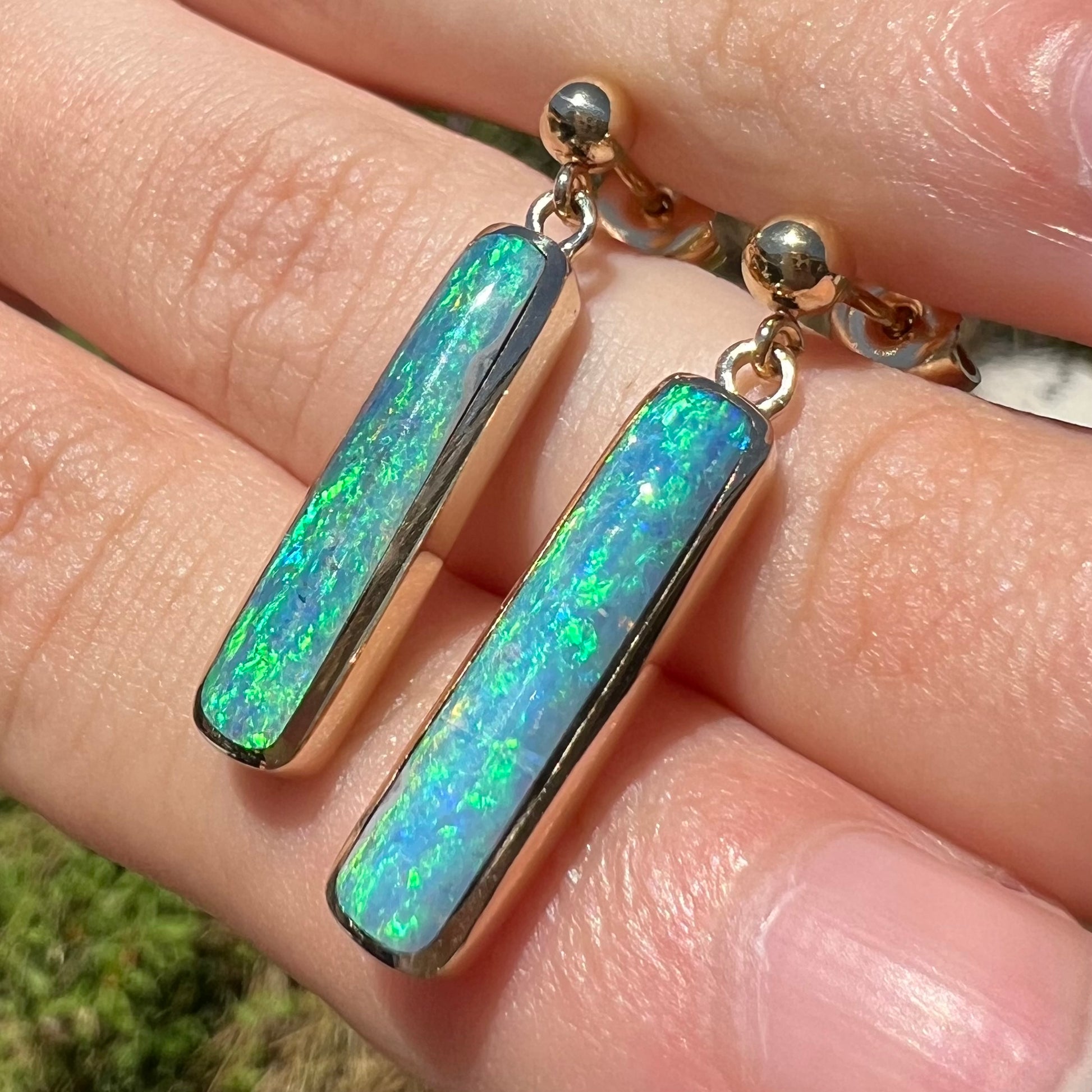 A pair of Lightning Ridge opal dangle earrings with green color play, shown in the sunlight at an angle.