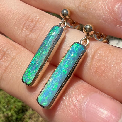 A pair of Lightning Ridge opal dangle earrings with green color play, shown in the sunlight at an angle.