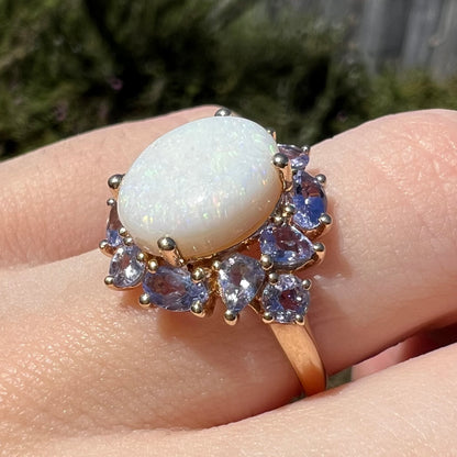 Angled side view of a lightweight 14k gold ring set with Australian opal and cluster of round, pear shaped, and oval tanzanite accents, worn on a finger in the sunlight