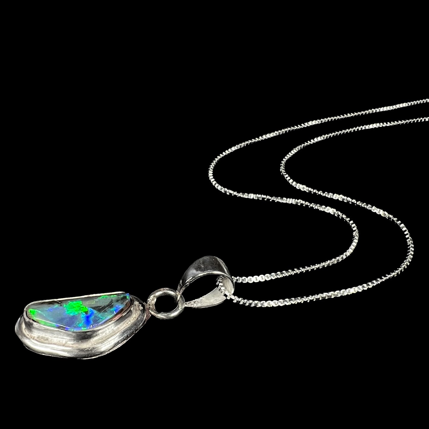 A sterling silver Australian black opal men's necklace shown from an angle laying on a black background, green and blue flashes visible in the opal.