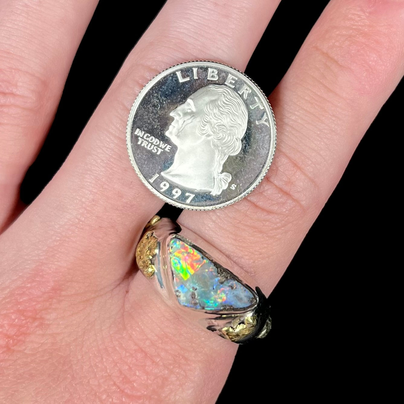 A men's Australian boulder opal solitaire ring shown close-up on a and next to a quarter coin.  The opal has green and red flashes of color.