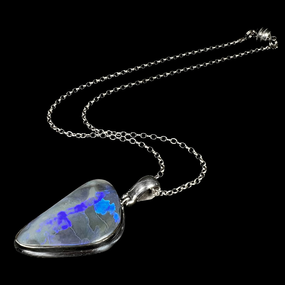 Angled view of men's Australian black opal necklace with blue flashes of color in sterling silver, shown on black background, its silver chain trailing behind the pendant