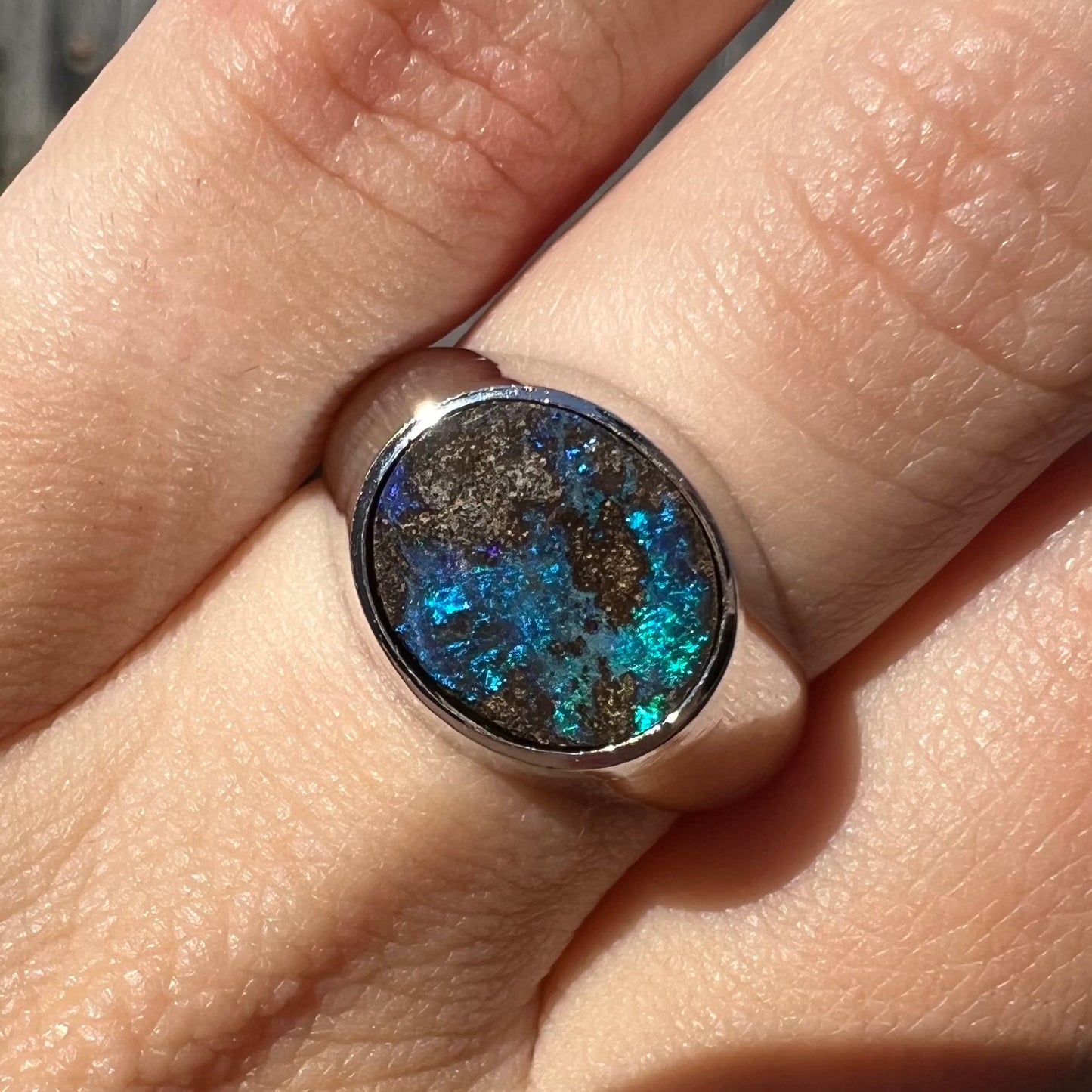 Close-up front view of a stainless steel men's Australian boulder opal ring with blue color play on a finger in sunlight.
