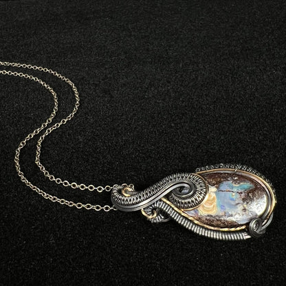 A men's Australian boulder opal wire wrapped necklace with blackened and gold-tone wire shown laying on a black velvet background, its chain flowing behind it.