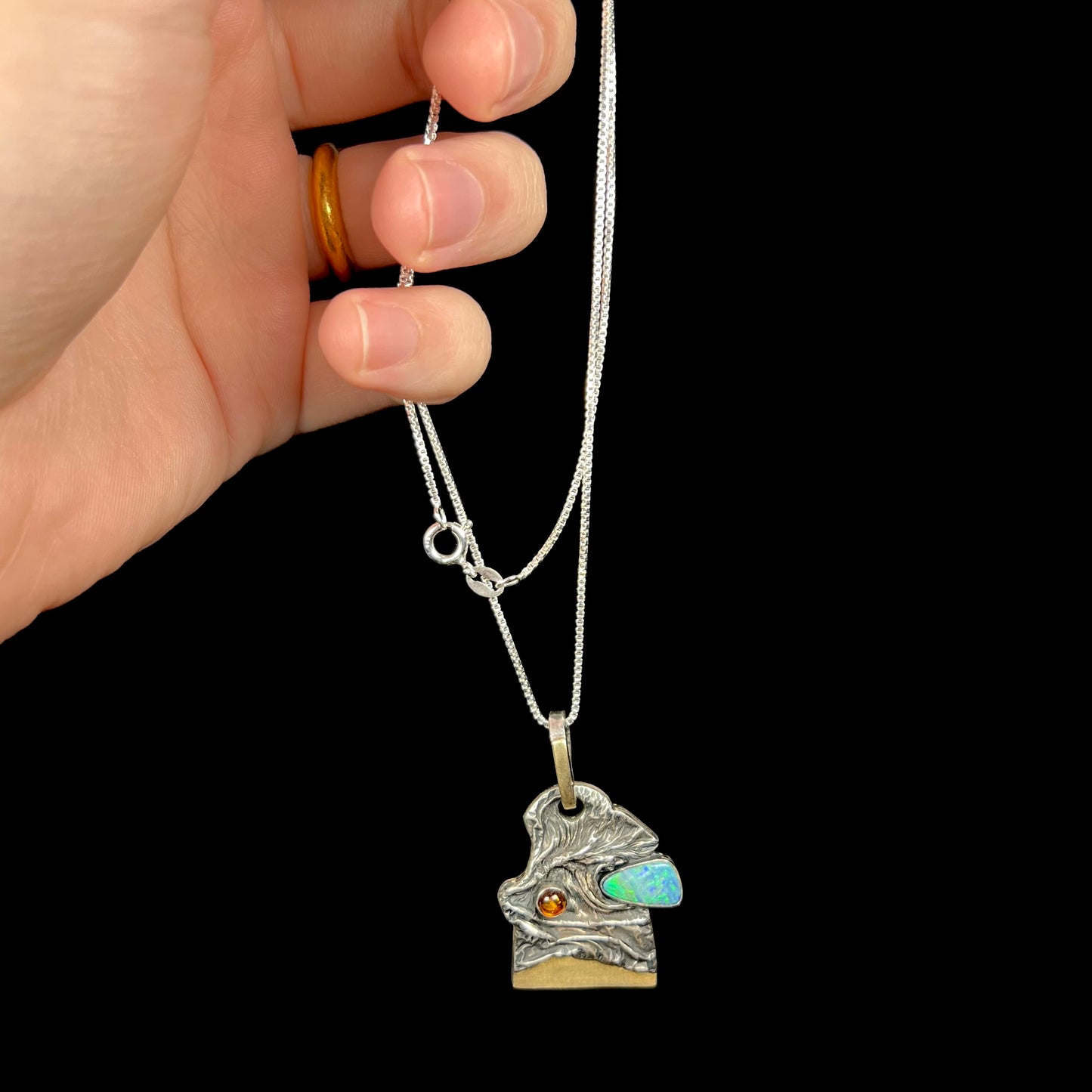 Hand dangling a men's brutalist-style sterling silver opal doublet and amber necklace from its silver box-style chain.