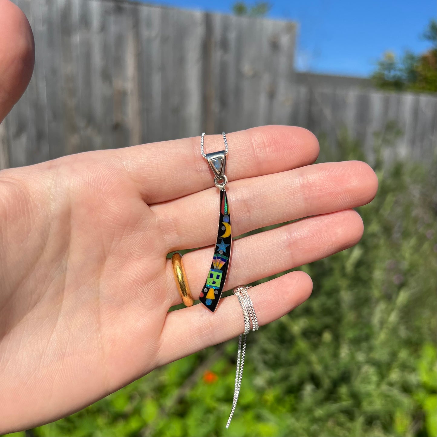 Hand holding men's elongated Southwest-style opal inlay pendant in sunlight against blurred outdoor background.